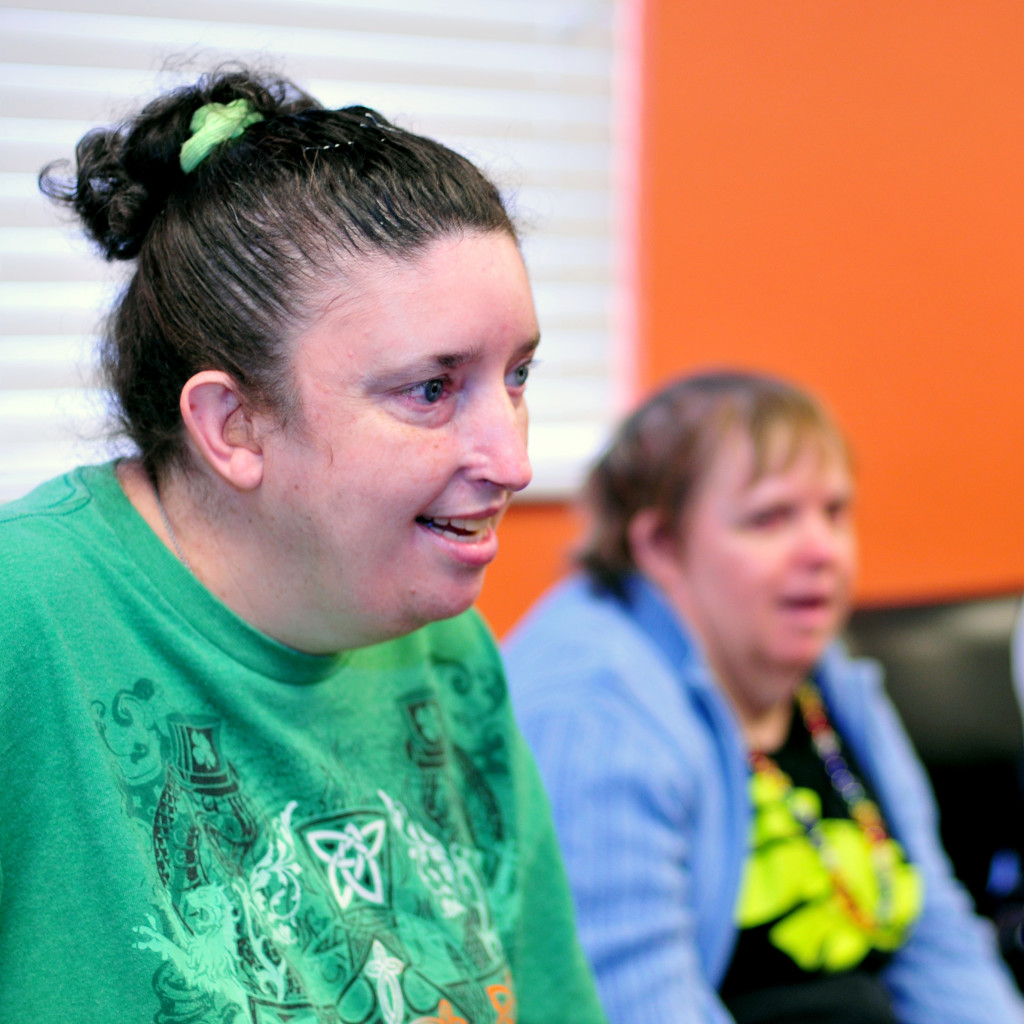 Woman in brown hair and a green shirt is grinning and sitting next to another woman on a couch